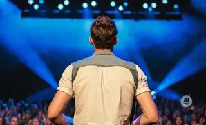 Man in a light-colored shirt with denim accents stands on a stage facing a cheering crowd under bright blue lights.