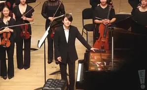 Pianist in a black suit stands by a grand piano, with an orchestra behind him.