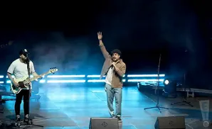 A male singer on stage with a microphone, right arm raised, beside a bass guitarist.