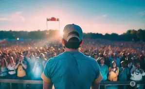 A person in a baseball cap and teal shirt stands on a stage, facing a large, cheering crowd at a concert during sunset.