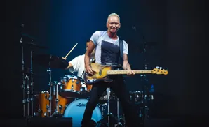 Bassist Sting smiles while playing a Fender bass guitar during a live performance, with a drummer in the background.