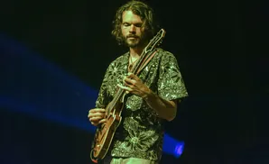 A man with long, dark, curly hair and a beard plays a guitar on stage, lit by blue and green stage lights.
