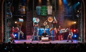 Performers on stage with buckets and brooms, some hitting metal drums.