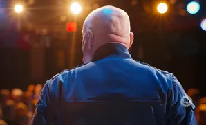 Back of a bald man in a dark blue uniform, facing away from the camera, with stage lights behind him.