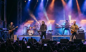 A band performs on stage with colorful lights and smoke, with a guitarist in the foreground and a crowd watching.