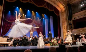 A ballet dancer leaps on stage during a performance with an orchestra, opera singer, and ballroom dancers.