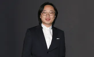 A man with glasses and long dark hair wearing a dark suit and white shirt smiles.