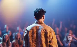 Man with curly hair in a patterned shirt faces a blurry, cheering crowd under blue and yellow stage lights.