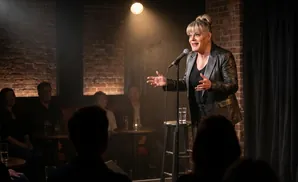 A comedian performs for a seated audience in a dimly lit brick room, illuminated by a spotlight.
