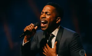 A Black man in a suit sings passionately into a microphone with his eyes closed and one hand on his chest.