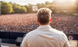Back of person in light shirt facing a large, blurred crowd at an outdoor concert with bright sunlight.