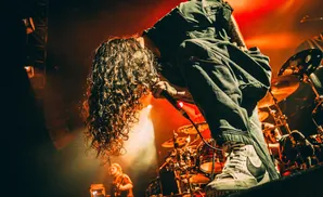 A rock singer with long, curly hair leans into a microphone on a stage lit with red and orange lights.