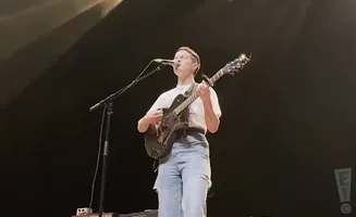 A person plays a guitar and sings into a microphone on a dark stage.