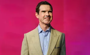 Man in a tan suit and blue shirt smiles against a bright pink background.