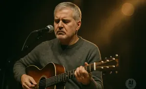 A man with grey hair plays an acoustic guitar into a microphone, bathed in warm stage lighting.