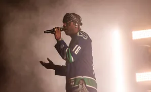 A rapper performs on stage wearing a blue and green hockey jersey, bandana, and jewelry.