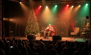A man reads a book while sitting in a red armchair on a stage next to a decorated Christmas tree, with colored spotlights overhead.