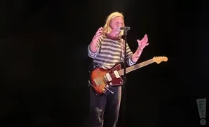 A male musician with long blonde hair plays an electric guitar and sings into a microphone on a dark stage.