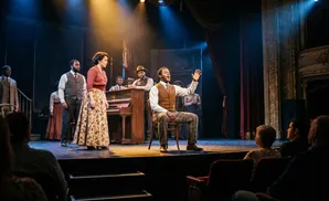 A Black man sings at a piano on stage with other performers.