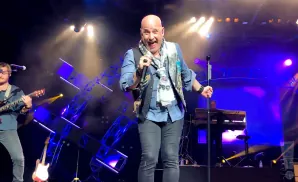A bald man in a blue shirt and vest sings into a microphone on a brightly lit stage.