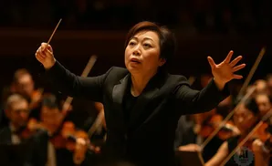 An Asian female orchestra conductor leads musicians, holding a baton and gesturing with her hands.