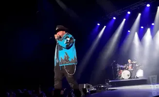 A male singer in a cowboy hat and bright blue jacket performs on stage with a drummer in the background.