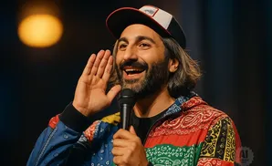 A man wearing a bandana-print jacket and a baseball cap tells a story into a microphone, cupping his ear.