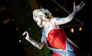 A tattooed woman in a red dress sings into a microphone on stage.