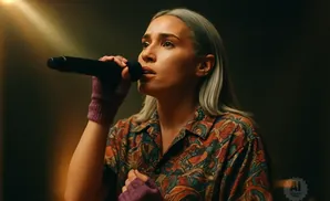 A woman with blonde hair sings into a microphone, wearing a colorful shirt and fingerless gloves.