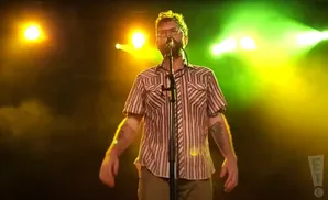 A man with glasses and a striped shirt stands on stage, singing into a microphone.