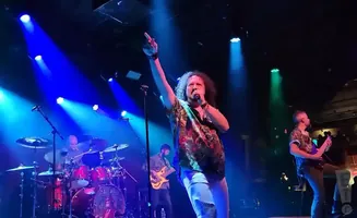 A rock band performs on a stage lit with blue and green lights. The lead singer, with curly hair, sings into a microphone.