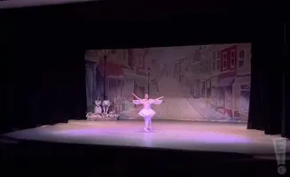 A ballerina in a white tutu performs on a stage with a painted backdrop of a street scene.