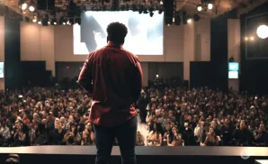 A speaker in a red shirt addresses a large, seated audience from a stage.