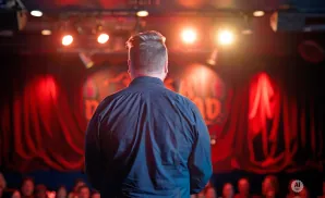 Man in a dark shirt stands on stage facing a red curtain and audience, lit by stage lights.