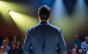 Man in a dark blue shirt facing away from the camera on a stage, with spotlights and an audience in the background.