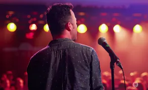 Man on stage facing away from camera, speaking into a microphone with colorful lights in the background.