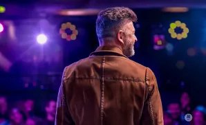 Man in a brown jacket seen from behind on a stage with purple lights and a blurred audience.