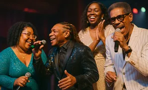A Black male singer in a sparkly black suit sings into a microphone on stage with two female singers.
