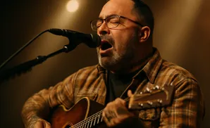 Man with glasses singing and playing an acoustic guitar, wearing a plaid shirt.
