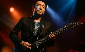 A male guitarist with a skull lapel pin plays a black electric guitar on stage under red and blue lights.