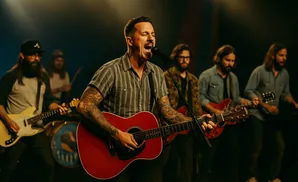 A tattooed man sings and plays a red guitar, backed by a band on a dimly lit stage.