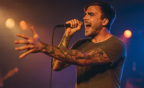 A tattooed man sings into a microphone on stage, bathed in warm stage lights.
