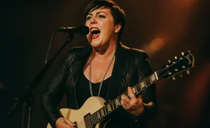 A woman with short hair sings into a microphone while playing a cream-colored electric guitar on stage.