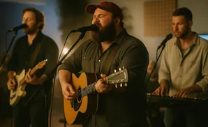A band of three men play music. The lead singer strums an acoustic guitar and sings into a microphone, with other musicians playing electric guitar and …