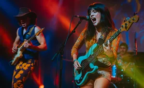 A woman sings and plays a bass guitar on stage, with a man playing guitar beside her.