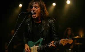A male singer with long hair, wearing a black leather jacket, sings into a microphone while playing a teal electric guitar on stage.