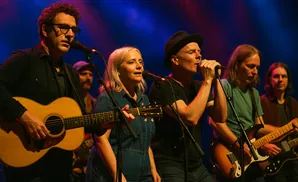 A band plays on a stage with blue and pink lighting. A guitarist in the foreground wears glasses and plays an acoustic guitar.