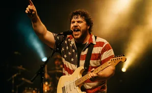 A male guitarist in an American flag shirt sings and plays a cream-colored electric guitar on stage with warm lighting.