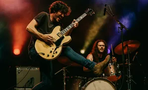 A guitarist performs on a cream-colored electric guitar, with a drummer in the background.