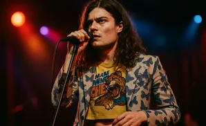 Male singer with long curly hair, wearing a butterfly-patterned jacket and a yellow t-shirt, performs into a microphone.